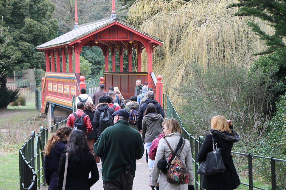 A group of people guided by a ranger walking toward the red Swiss Bridge in Birkenhead Park