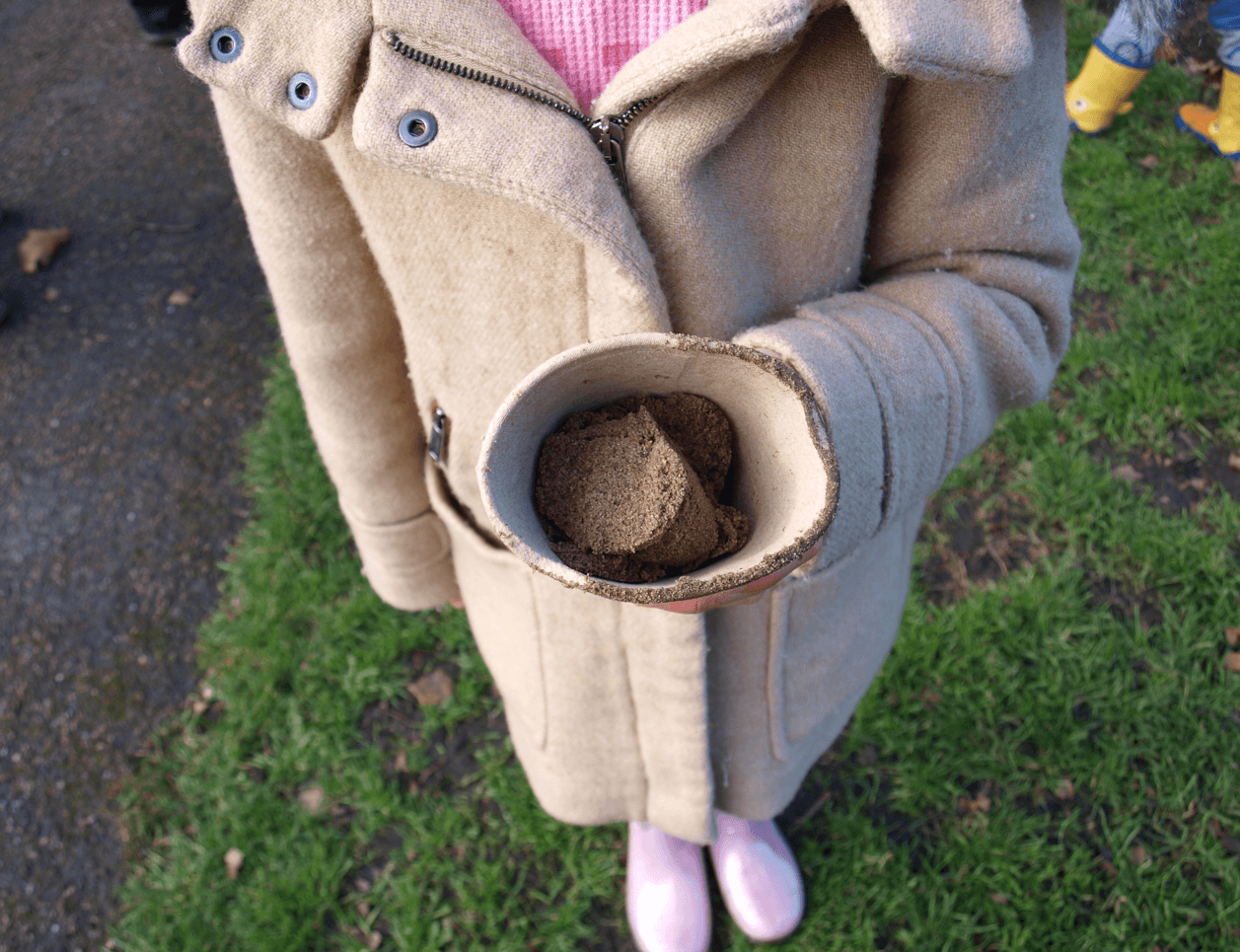 A little girl holding a cardboard pot with a mixture of soil and seeds in it